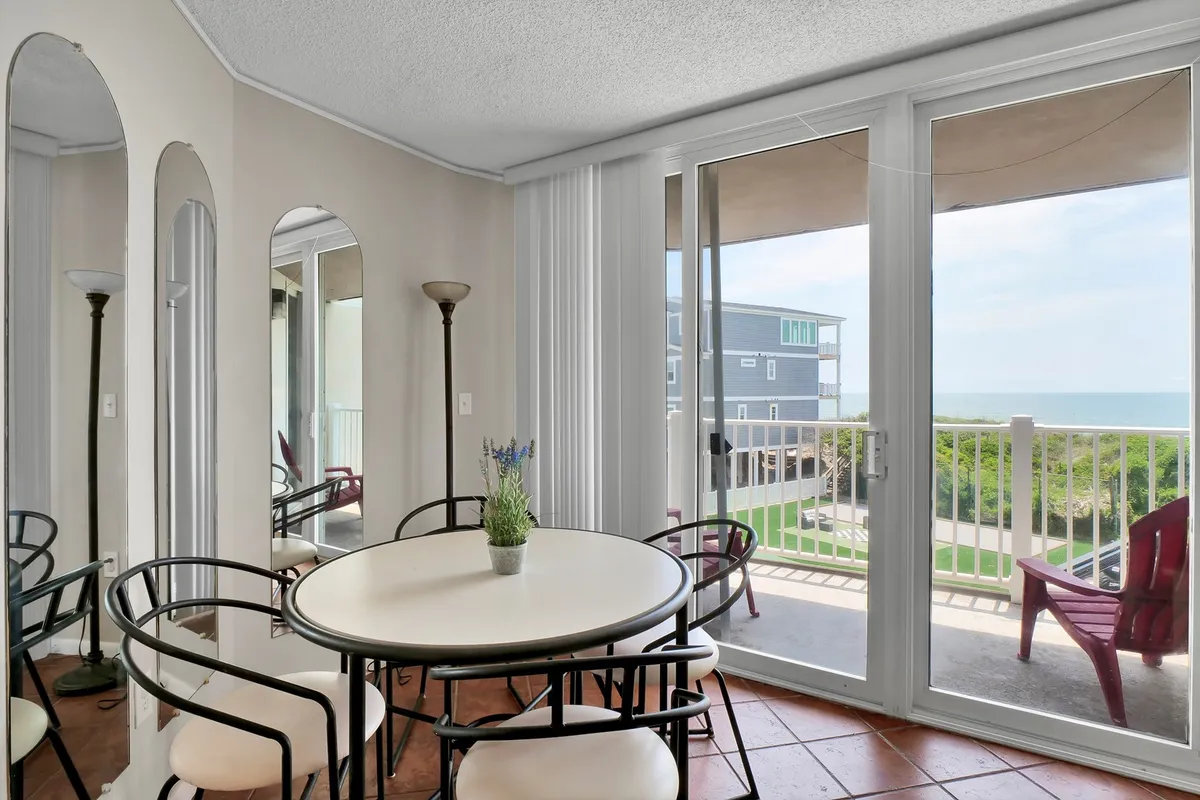 A cheerful dining space with a round table set beside large windows, offering unobstructed ocean views. The room is bright and inviting, with a decorative plant and mirrors enhancing the sense of openness and connection to the sea.