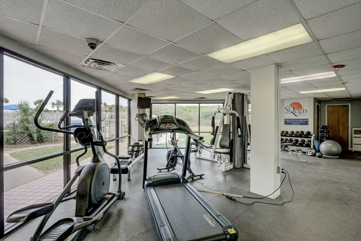 A bright fitness center with large windows, featuring a treadmill, elliptical, and weight equipment. The inviting space encourages wellness activities and offers an uplifting start or end to the day, in line with the wellness-focused amenities described in the section.