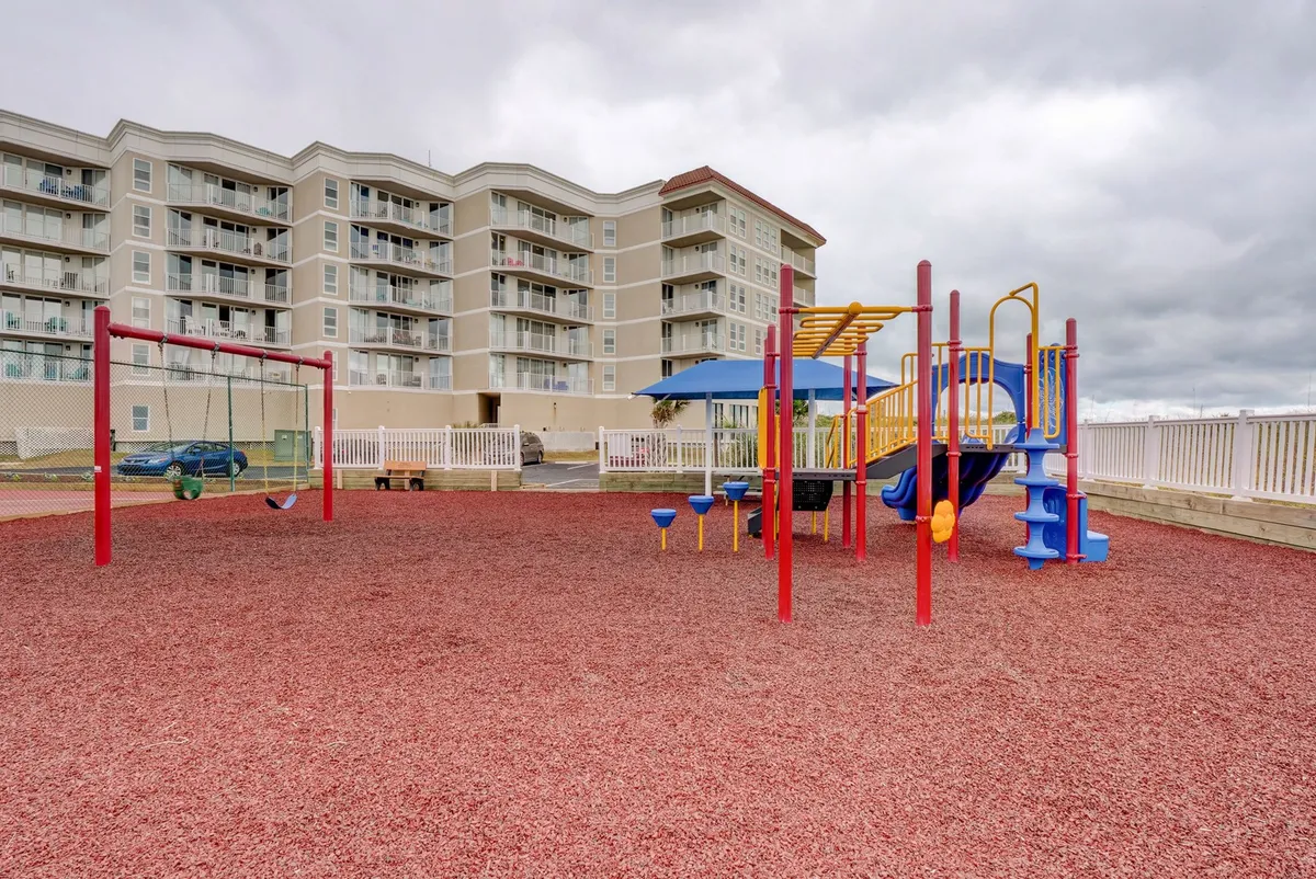 A vibrant playground featuring a variety of colorful equipment and swings, set on a safe red rubber surface. Children are seen playing and families enjoy the energetic, joyful atmosphere. The scene evokes laughter, family bonding, and active outdoor fun just steps from the condo.