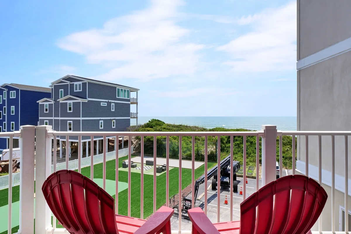 The image showcases a tranquil balcony setting overlooking the sparkling ocean, with vibrant red chairs inviting guests to relax. Lush green lawn stretches below, while a wide blue sky completes the peaceful, open-air scene. The image evokes a sense of relaxation, inviting viewers to imagine themselves savoring coffee, ocean breezes, and moments with loved ones.