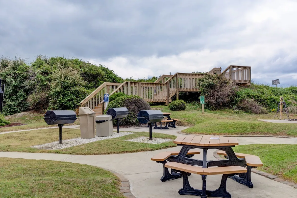 A casual outdoor grilling and picnic area with wooden picnic tables and multiple charcoal grill stations. The relaxed setup encourages outdoor dining, gatherings, and classic beach barbecues after a day in the sun.