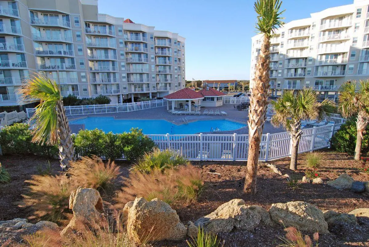 A sun-drenched outdoor swimming pool surrounded by palm trees and residential buildings. The pool area features ample space for lounging, swimming, and socializing, creating a true resort atmosphere.